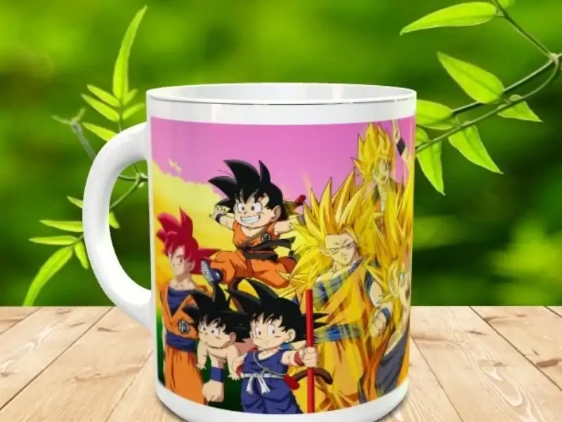 Taza de cerámica blanca con ilustración colorida de varios personajes del universo anime sobre una mesa de madera y fondo vegetal difuminado. Ref. goku-15x