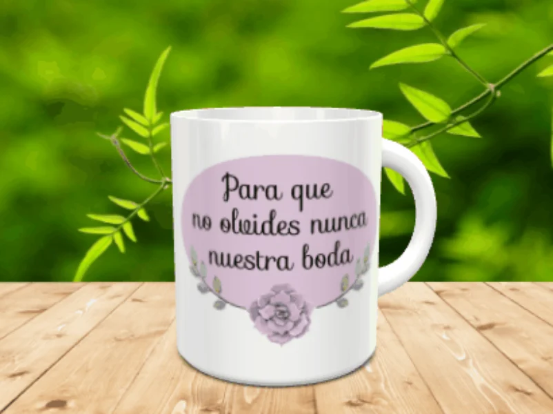 Maqueta de taza blanca de cerámica con un óvalo lila, la frase Para que no olvides nunca nuestra boda y un detalle floral sobre una mesa de madera.
