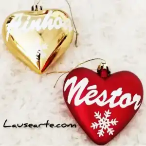 Fotografía real de dos adornos de Navidad personalizados con forma de corazón sobre fondo blanco. Incluye un corazón dorado brillante con el nombre Ainhoa y un corazón rojo con el nombre Néstor.