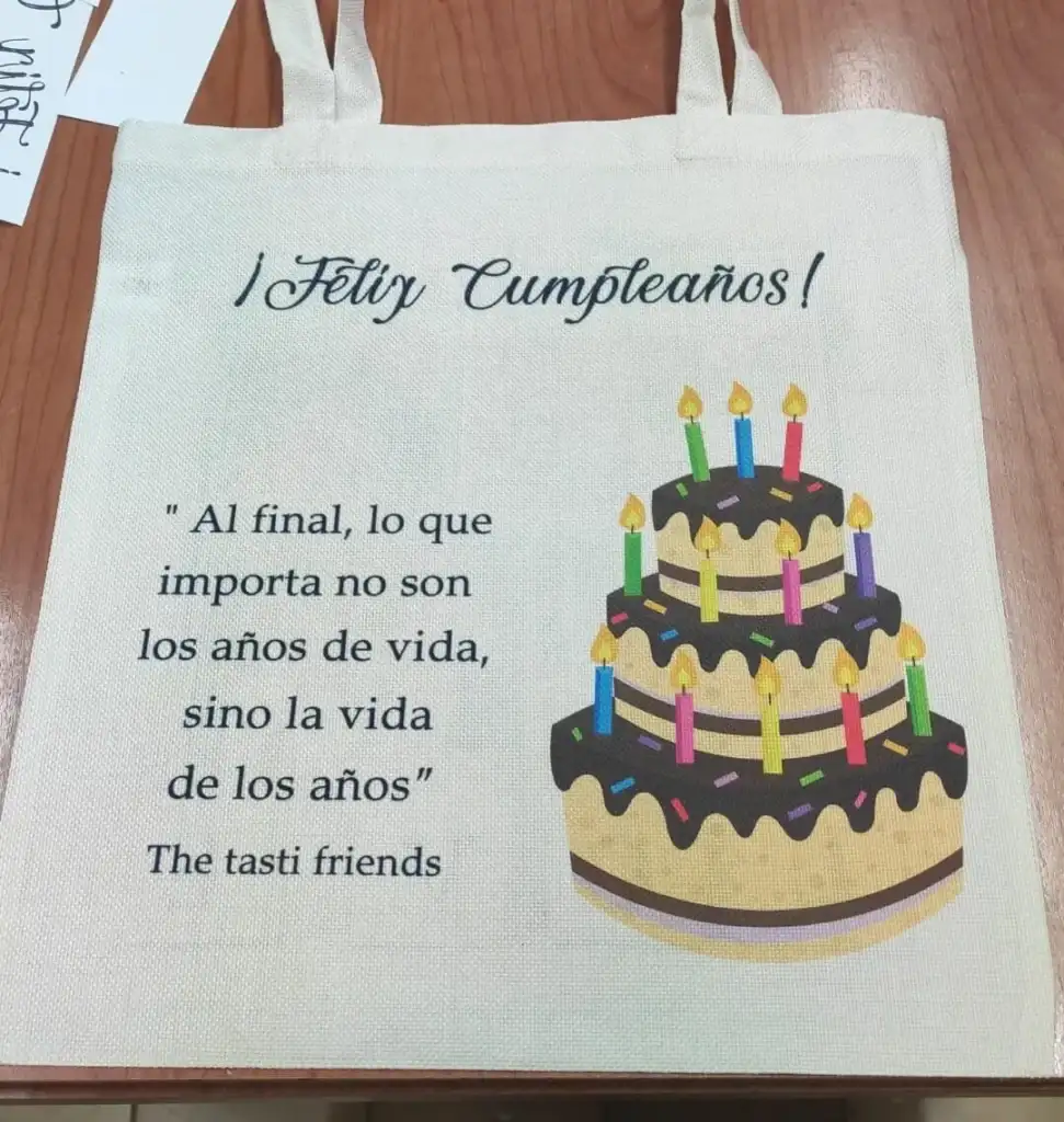 Fotografía de catálogo de una bolsa de tela de algodón tipo tote bag de color crudo. Diseño impreso de Feliz Cumpleaños con una tarta con velas, una frase célebre emotiva y dedicatoria de amigos.
