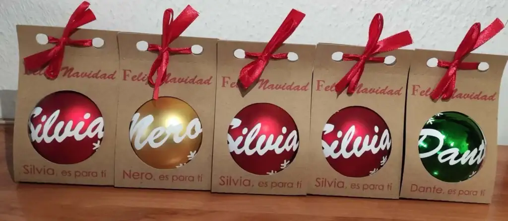 Fotografía real de cinco bolas de Navidad personalizadas a mano en varios colores: rojo, dorado y verde. Cada esfera lleva un nombre escrito en relieve y purpurina, presentadas en bolsas individuales kraft con lazo.