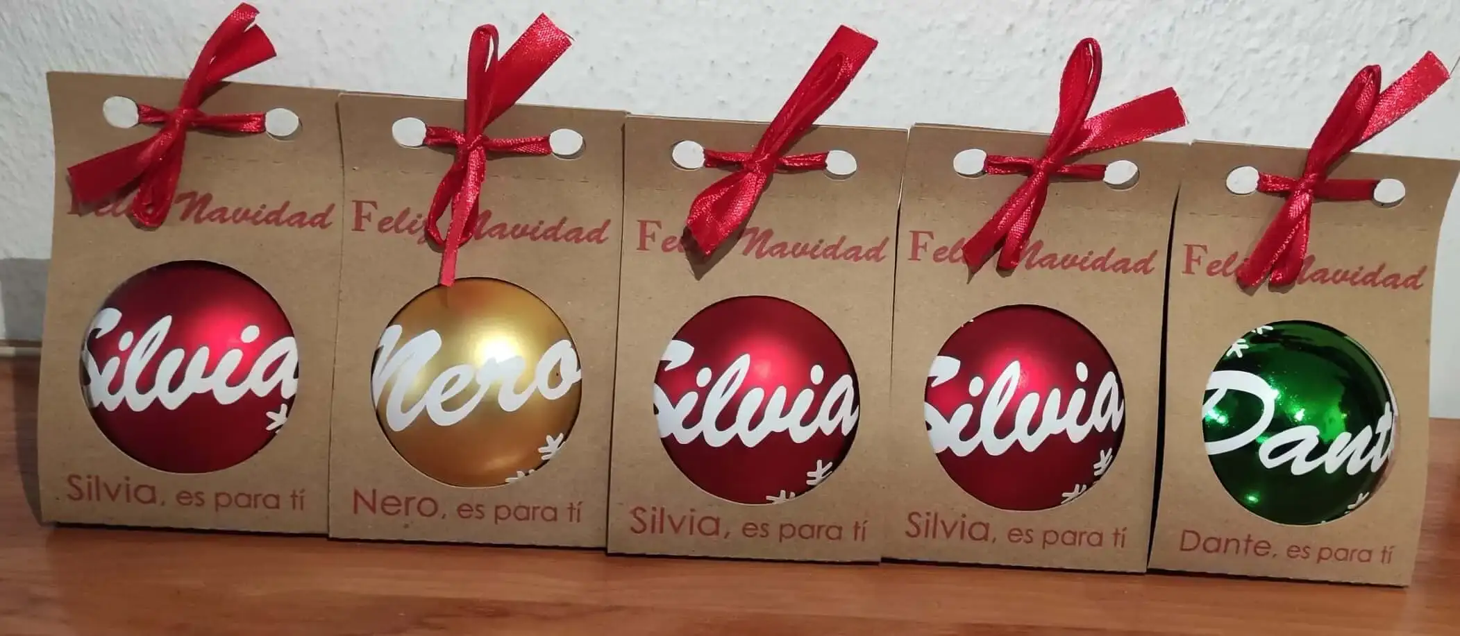 Fotografía real de cinco bolas de Navidad personalizadas a mano en varios colores: rojo, dorado y verde. Cada esfera lleva un nombre escrito en relieve y purpurina, presentadas en bolsas individuales kraft con lazo.