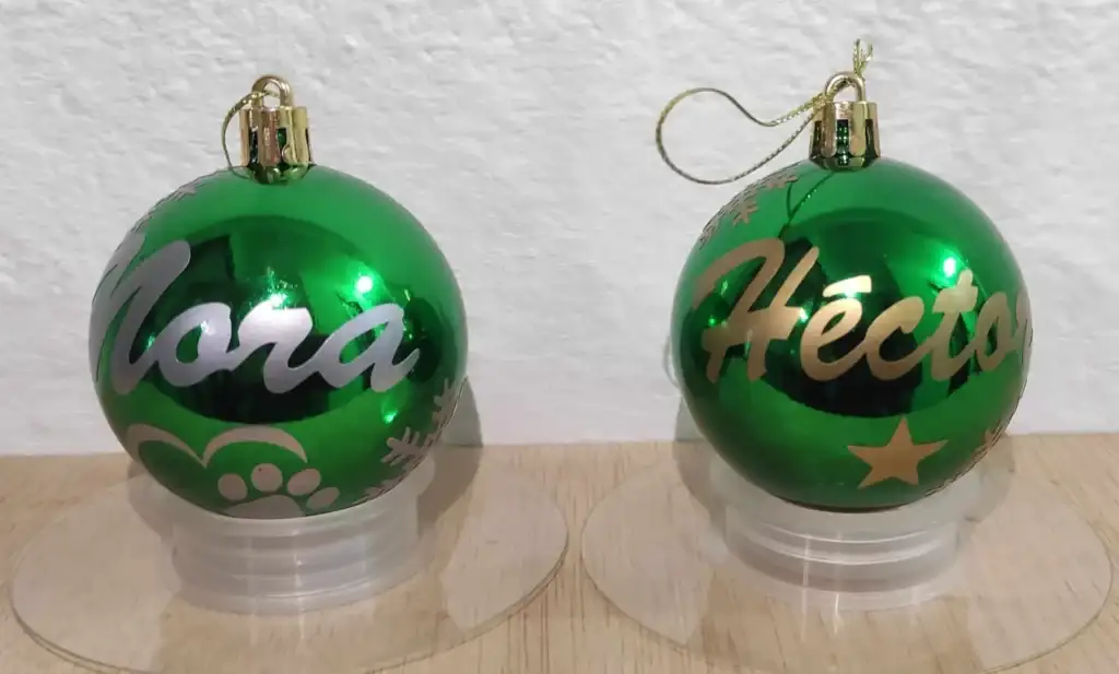 Fotografía real de dos bolas de Navidad verdes brillantes sobre soportes. Esferas navideñas personalizadas a mano con purpurina con los nombres de Cora y Héctor, decoradas con copos y estrellas.