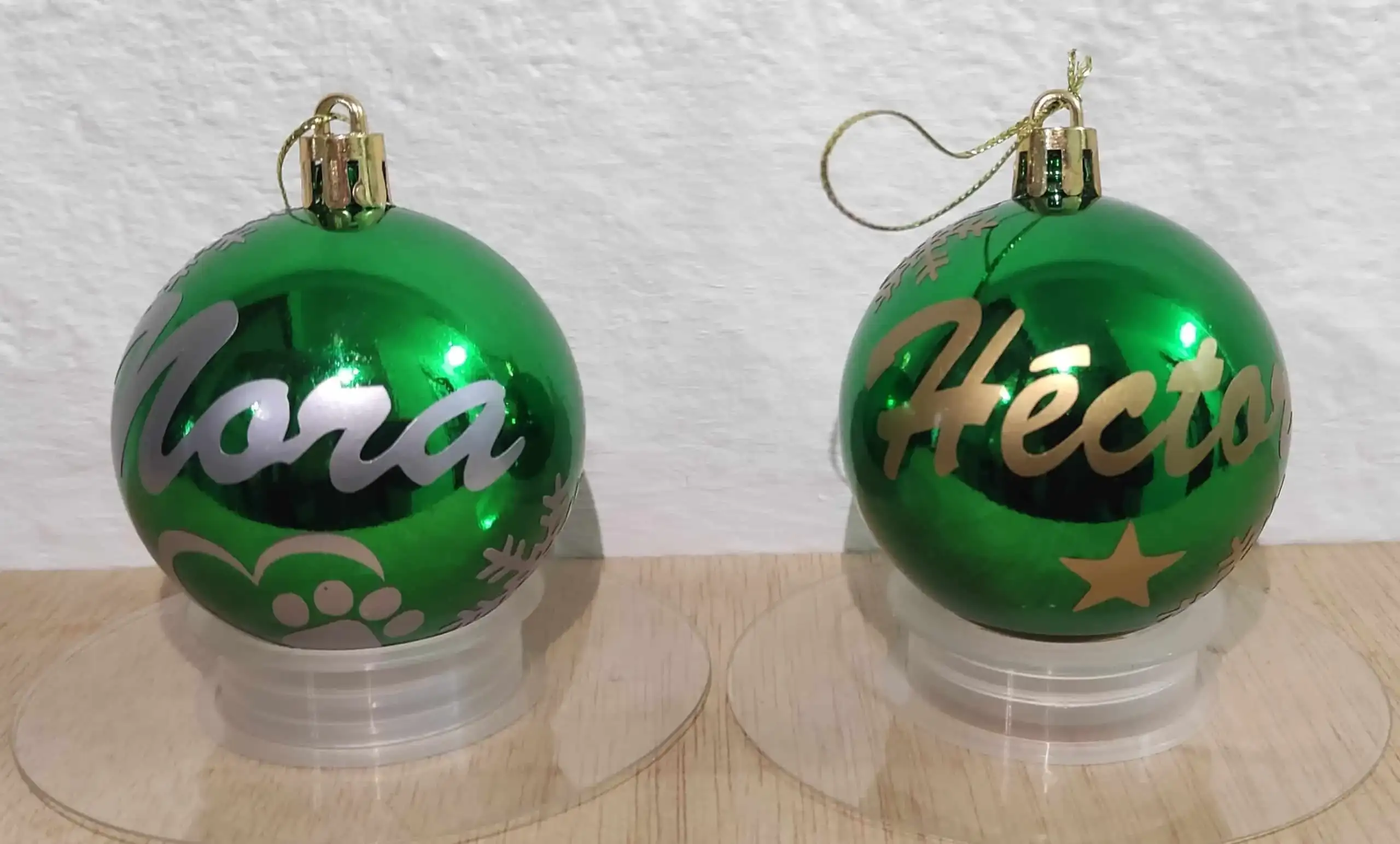 Fotografía real de dos bolas de Navidad verdes brillantes sobre soportes. Esferas navideñas personalizadas a mano con purpurina con los nombres de Cora y Héctor, decoradas con copos y estrellas.