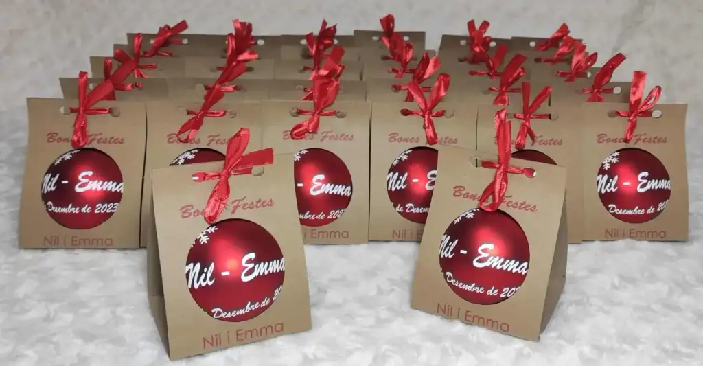 Fotografía real de un lote de regalos navideños para colegios en bolsas kraft individuales con lazo rojo, acompañadas de bolas de Navidad rojas personalizadas con nombres de alumnos.