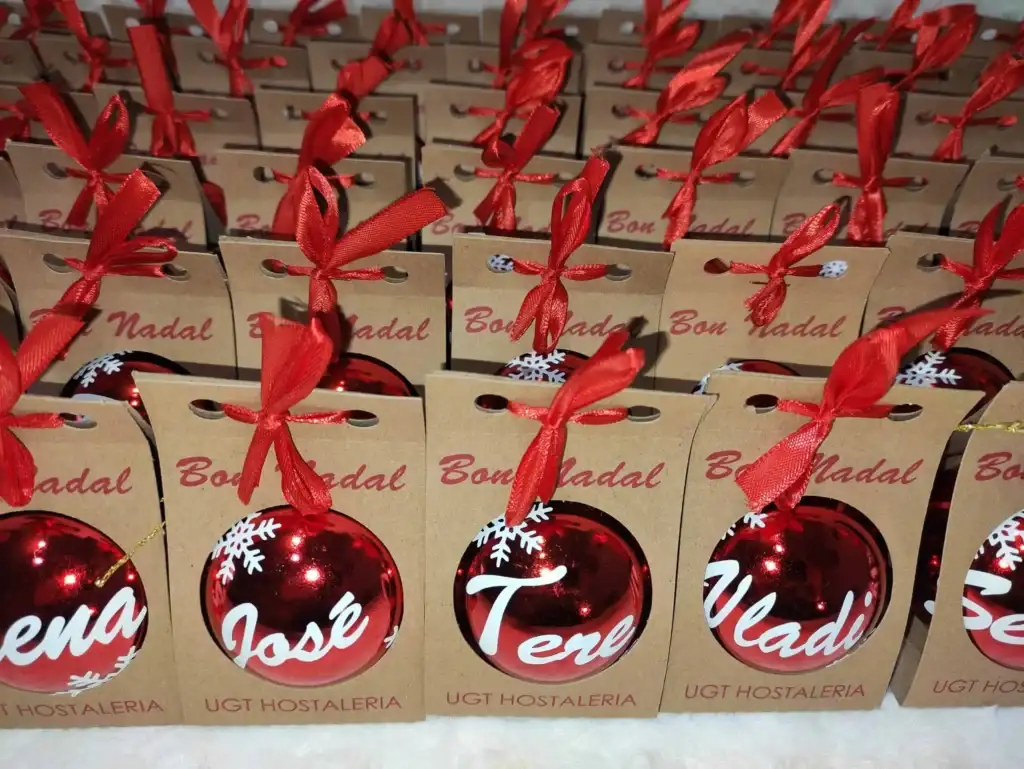 Fotografía real de un lote masivo de regalos de Navidad corporativos. Muestra bolsas de cartón kraft con el mensaje Bon Nadal y lazo rojo, junto a bolas navideñas rojas personalizadas con nombres y copos de nieve.