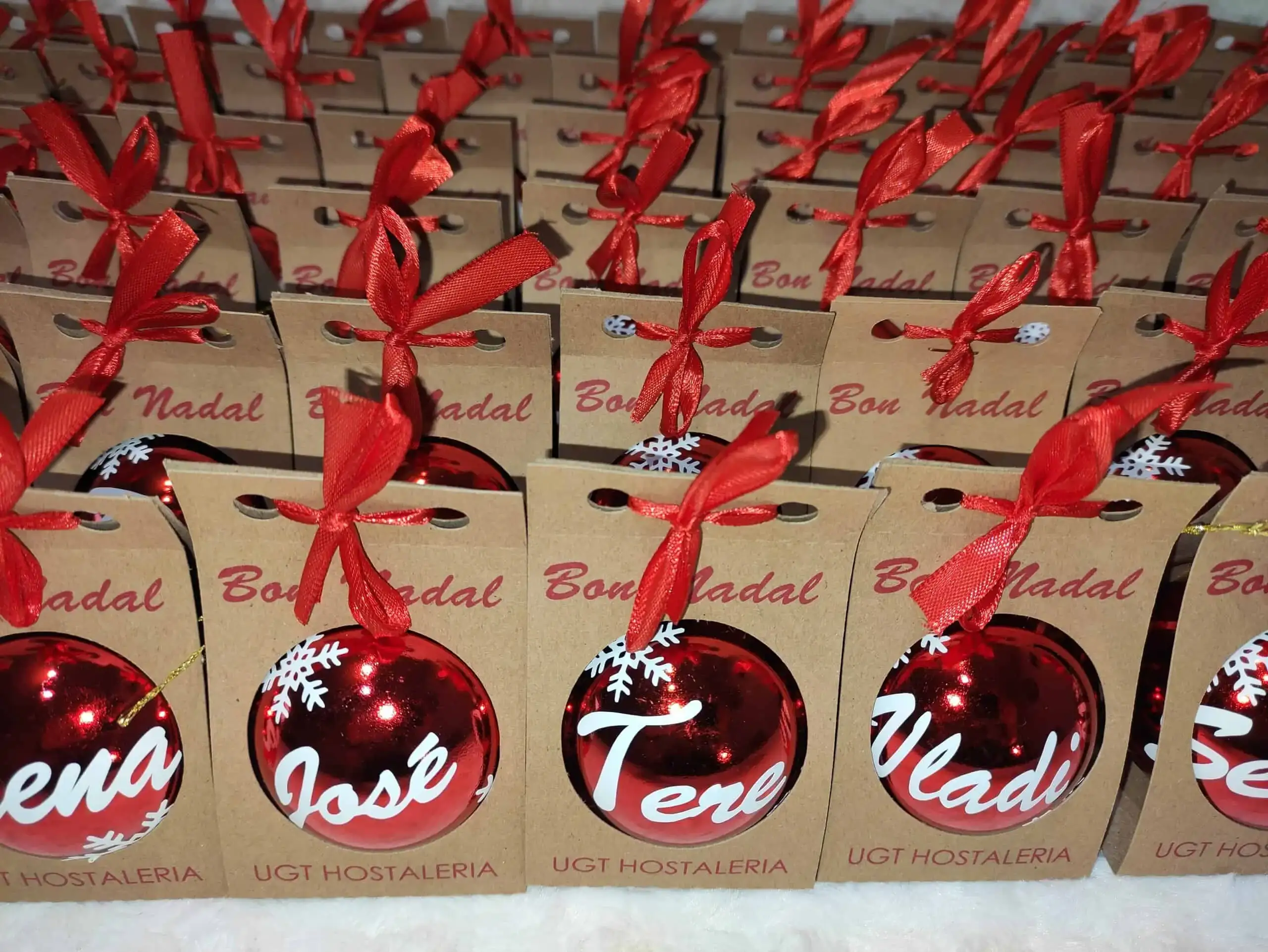 Fotografía real de un lote masivo de regalos de Navidad corporativos. Muestra bolsas de cartón kraft con el mensaje Bon Nadal y lazo rojo, junto a bolas navideñas rojas personalizadas con nombres y copos de nieve.
