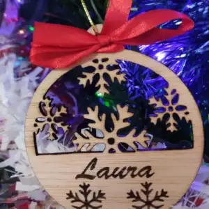Fotografía en primer plano de un adorno navideño de madera colgado en el árbol. Bola de Navidad personalizada de madera natural calada con copos de nieve y el nombre Laura grabado con láser, decorada con un lazo rojo.