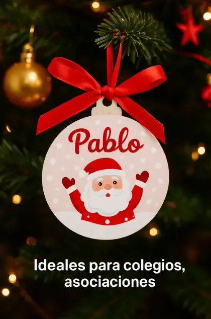 Adorno navideño blanco infantil colgado en las ramas de un árbol de Navidad iluminado. Está personalizado con el nombre Pablo en letras rojas y un dibujo de Papá Noel. Un texto superpuesto indica: Ideales para colegios, asociaciones.
