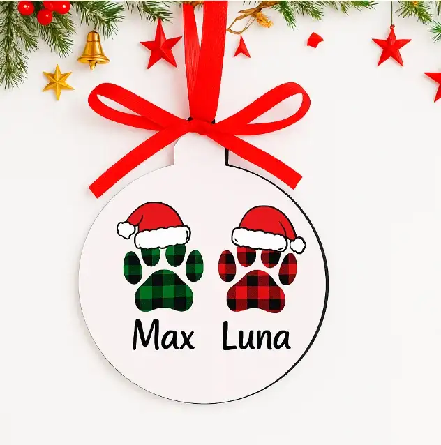 Adorno navideño blanco personalizado con los nombres Max y Luna debajo de dos huellas de mascota. Las huellas tienen estampado de cuadros escoceses (verde y rojo) y gorritos de Papá Noel. Incluye un lazo rojo para colgar.