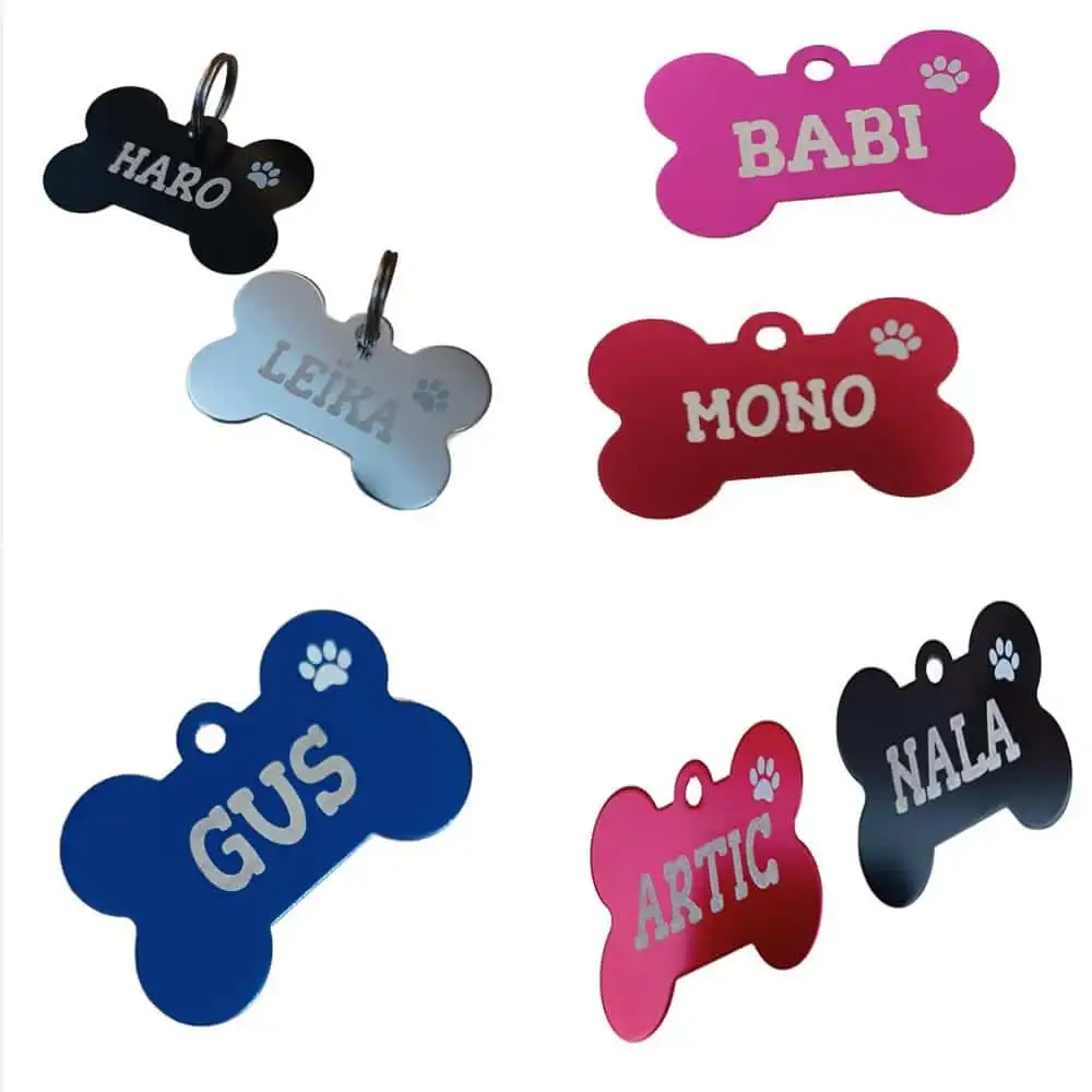 Collage de catálogo sobre fondo blanco que muestra una colección de placas identificativas de aluminio para perros en forma de hueso. Se aprecian varios colores y modelos grabados con nombres como Gus, Nala y Haro junto a huellas caladas.