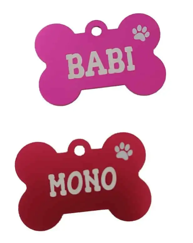 Fotografía de catálogo sobre fondo blanco de dos placas para perros con forma de hueso grabadas con láser. La chapa de arriba es fucsia con el nombre Babi y la de abajo es roja con el nombre Mono, ambas con el icono de una huella calada.