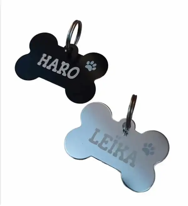 Fotografía de catálogo en perspectiva sobre fondo blanco de dos placas metálicas para perros con forma de hueso y argolla incluida. Se muestra un modelo negro grabado con el nombre Haro y un modelo plata con el nombre Leika, ambos con una huella calada.
