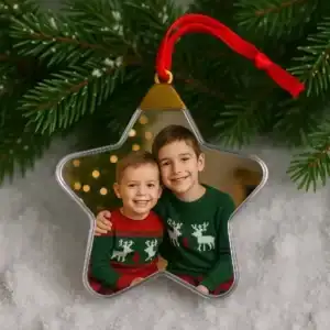 Adorno para el árbol de Navidad con forma de estrella transparente, personalizado con la fotografía de dos hermanos pequeños sonriendo con jerseys navideños de renos. Colgado de una rama con luces de fondo.