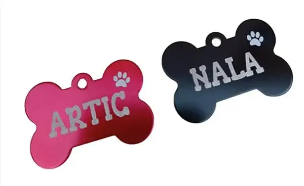 Fotografía de catálogo sobre fondo blanco que muestra en perspectiva dos placas para perros en forma de hueso. A la izquierda se ve un modelo fucsia grabado con el nombre Artic, y a la derecha un modelo negro mate con el nombre Nala, ambos con una huella calada.