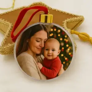 Bola de Navidad esférica y transparente con colgador dorado y cinta de terciopelo rojo. En el interior muestra una fotografía personalizada de una madre abrazando con ternura a su bebé frente al árbol iluminado.
