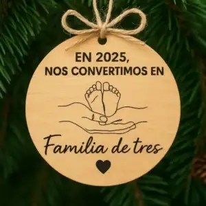Adorno navideño de madera grabado con frase En 2025 nos convertimos en familia de tres y dibujo de pies de bebé.